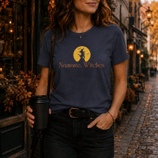 A woman wearing a navy blue graphic t-shirt with a gold witch silhouette on a full moon and the text 'Namaste, Witches.' She is holding a black coffee cup on a cobblestone street at night.