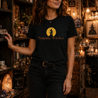 A woman wearing a black graphic t-shirt with a yellow moon and witch silhouette and the text 'Namaste, Witches' printed on the front, standing in a cozy, dimly lit shop filled with crystals and candles.