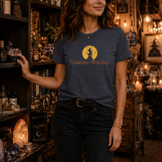 A woman wearing a navy blue graphic t-shirt with a gold witch and moon design and the text 'Namaste, Witches' printed on the front, standing in a cozy, dimly lit shop filled with crystals and books.