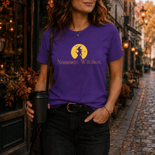 A woman wearing a purple graphic t-shirt with a yellow moon and witch silhouette and the text 'Namaste, Witches.' standing in a cobblestone street.