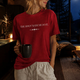 A woman wearing a red graphic t-shirt with the text 'THE MOON MADE ME DO IT.' in white, holding a black mug in a cozy, dimly lit home interior.