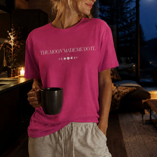 A woman wearing a bright pink t-shirt with the white text 'THE MOON MADE ME DO IT.' and moon phase graphic, holding a black mug in a cozy, dimly lit bedroom setting.