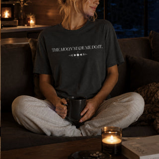 A woman sits cross-legged on a dark couch in a cozy living room, wearing a dark charcoal t-shirt with the white text 'THE MOON MADE ME DO IT.' and a moon phase graphic, paired with light gray sweatpants and holding a black mug.