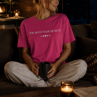 A woman sits cross-legged on a dark couch in a cozy room, wearing a bright pink graphic T-shirt that reads 'THE MOODY MADE ME DO IT.' with moon phase icons, and light gray sweatpants while holding a black mug.