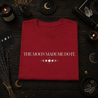A folded red t-shirt with white text reading 'THE MOON MADE ME DO IT.' and a moon phase graphic, displayed on a dark wooden surface surrounded by occult-themed props like candles, crystals, and a journal.