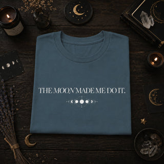 A folded slate blue t-shirt with the white text 'THE MOON MADE ME DO IT.' and a moon phase graphic, displayed in a flat-lay on a dark wood background surrounded by candles, crystals, and moon phase cards.