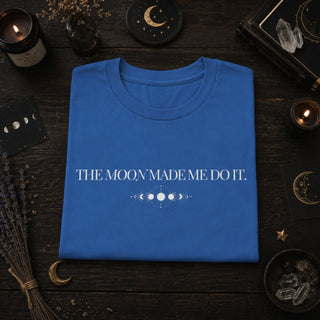 A folded blue T-shirt with white text reading 'THE MOON MADE ME DO IT.' and a moon phase graphic, displayed on a dark wooden surface surrounded by candles, crystals, and moon-themed decor.