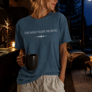 A woman wearing a slate blue graphic t-shirt that reads 'THE MOOY MADE ME DO IT.' in white text, holding a black mug in a cozy, dimly lit home interior.