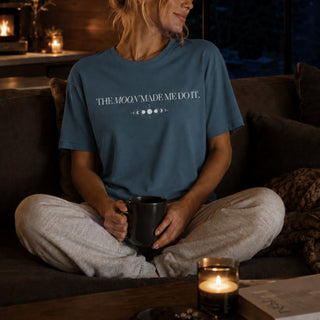 A woman sits cross-legged on a dark couch in a cozy room, wearing a slate blue graphic t-shirt with the white text 'THE, MOOY MADE ME DO IT.' and light heather gray sweatpants, holding a black mug.