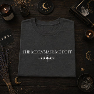 A folded dark heather gray t-shirt with white text reading 'THE MOON MADE ME DO IT.' and a moon phase graphic, displayed in a flat-lay on a dark wood surface surrounded by candles, crystals, dried lavender, and moon-themed cards.