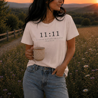 A woman standing in a field at sunset wearing a light beige graphic T-shirt with the text '11:11 IF YOU'RE WAITING FOR A SIGN, THIS IS IT.' and light blue jeans, holding a speckled ceramic mug.