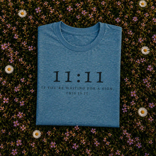 A folded heather blue t-shirt with black printed text reading '11:11 IF YOU'RE WAITING FOR A SIGN THIS IS IT.' is laid flat on a bed of small pink and white flowers.