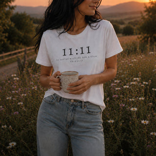 A woman outdoors in a field at sunset wearing a white graphic T-shirt with the text '11:11 IF YOU'RE WAITING FOR A SIGN, THIS IS IT.' and light-wash jeans, holding a speckled mug.