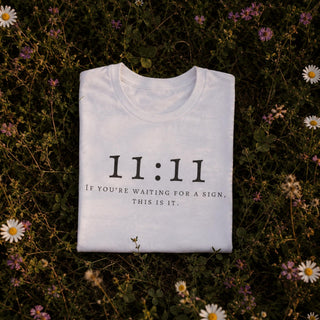 A folded white t-shirt with black text reading '11:11 IF YOU'RE WAITING FOR A SIGN, THIS IS IT.' is laid flat on a bed of green grass and small white daisies.