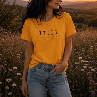 A woman wearing a mustard yellow graphic t-shirt with the text '11:11 IF YOU'RE WAITING FOR A SIGN, THIS IS IT.' printed in black, paired with light-wash blue jeans, standing in a field of wildflowers at sunset.