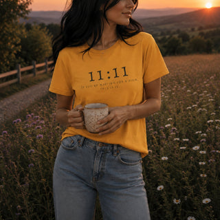 A woman standing outdoors in a field at sunset wearing a mustard yellow graphic t-shirt with the text '11:11 IF YOU ARE WAITING FOR A SIGN, WAIT FOR IT.' She is holding a speckled ceramic mug.