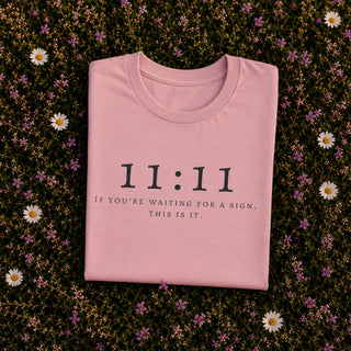 A folded light pink t-shirt with black printed text reading '11:11 IF YOU'RE WAITING FOR A SIGN, THIS IS IT.' is laid flat on a bed of small white daisies and purple flowers.