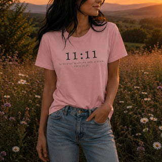 A woman wearing a light pink graphic t-shirt with the text '11:11 IF YOU'RE WAITING FOR A SIGN, THIS IS IT.' in black, paired with light-wash blue jeans, standing in a field of wildflowers at sunset.