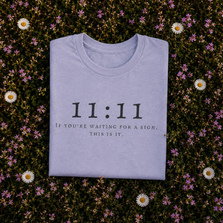 A folded light lavender t-shirt with black text reading '11:11 IF YOU'RE WAITING FOR A SIGN, THIS IS IT.' is laid flat on a bed of green foliage and small white and pink flowers.