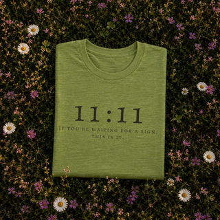 A folded sage green t-shirt with black text reading '11:11 IF YOU'RE WAITING FOR A SIGN, THIS IS IT.' is laid flat on a bed of green foliage and small white and purple flowers.