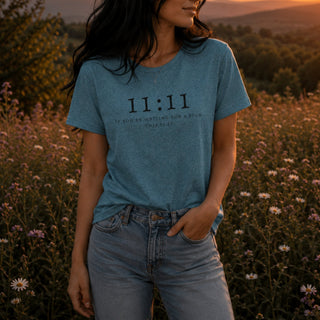 A woman wearing a heathered blue t-shirt with black text reading '11:11 IF YOU'RE WAITING FOR A SIGN, THIS IS IT' stands in a field of wildflowers at sunset.