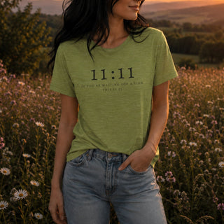 A woman wearing a light olive green heathered t-shirt with '11:11 IF YOU ARE WAITING FOR A SIGN, THINGS ARE IT' printed in black text, standing in a field of wildflowers at sunset.