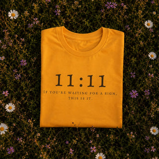 A folded mustard yellow t-shirt with black text reading '11:11 IF YOU'RE WAITING FOR A SIGN, THIS IS IT.' is laid flat on a bed of green grass and small white and purple wildflowers.