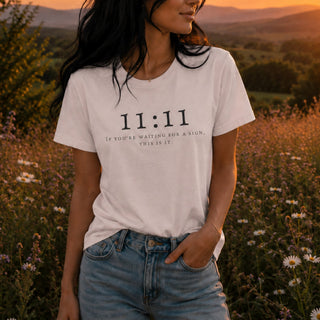 A woman wearing a white graphic t-shirt with the text '11:11 IF YOU'RE WAITING FOR A SIGN, THIS IS IT.' in black print, paired with light-wash blue jeans, standing in a field of wildflowers during a warm sunset.