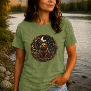 A woman wearing a heather sage green t-shirt with a circular graphic of a bear under a crescent moon, standing by a river at sunset.