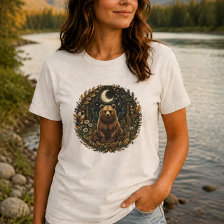 A woman wearing a light beige crewneck t-shirt with a circular graphic of a bear under a crescent moon in a forest, standing by a river.