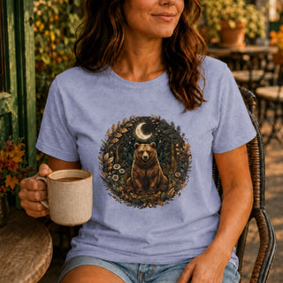 A woman wearing a light periwinkle blue graphic t-shirt with a circular forest bear design, sitting in a wicker chair and holding a mug on an outdoor patio.