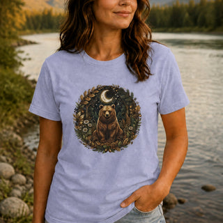 A woman wearing a light periwinkle blue t-shirt with a circular graphic of a brown bear under a crescent moon surrounded by foliage, standing by a river.