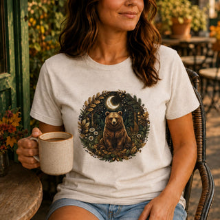 A woman sitting outdoors on a wicker chair wearing a light beige graphic t-shirt with a circular forest illustration of a brown bear under a crescent moon, holding a ceramic mug.