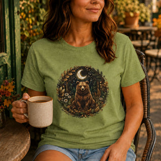 A woman sitting outdoors on a wicker chair wearing a sage green graphic t-shirt with a circular design of a brown bear under a crescent moon in a forest. She is holding a beige ceramic mug.
