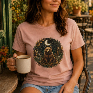 A woman sitting outdoors in a wicker chair wearing a dusty pink t-shirt with a circular graphic of a brown bear under a crescent moon, holding a mug.