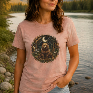 A woman wearing a light pink t-shirt with a circular graphic of a brown bear under a crescent moon in a forest, standing by a river.