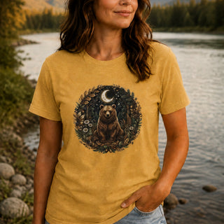 A woman wearing a mustard yellow graphic t-shirt with a circular print of a bear under a crescent moon, standing by a river with trees in the background.