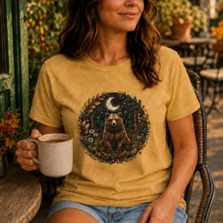 A woman sitting outdoors on a patio chair wearing a mustard yellow graphic t-shirt with a circular print of a bear under a crescent moon, holding a ceramic mug.