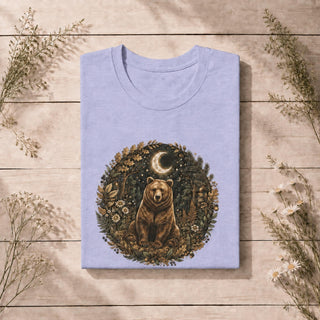 A folded light lavender crewneck t-shirt with a circular graphic of a brown bear in a forest under a crescent moon, displayed on a rustic wooden background with dried flowers.