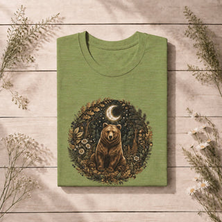 A folded sage green t-shirt with a detailed graphic of a bear in a forest circle, displayed on a light wood plank background with dried floral accents.