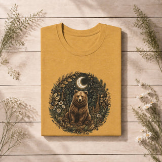 A folded mustard yellow t-shirt with a detailed graphic of a brown bear in a forest circle, displayed on a light wood plank background with decorative wildflowers.