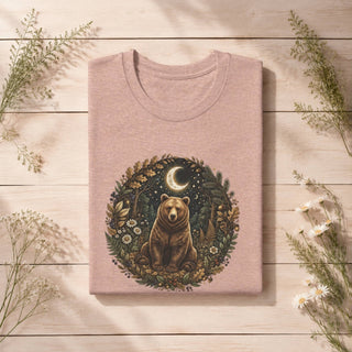 A folded dusty pink t-shirt with a detailed graphic of a bear in a forest circle print, displayed on a light wood plank background with decorative wildflowers.