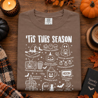 A brown crewneck t-shirt with a white 'Halloween Season' graphic print, laid flat on a rustic wooden table surrounded by seasonal props like a candle, pumpkin, and fall leaves.