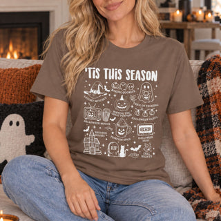 A woman sitting on a couch wearing a brown graphic t-shirt with white text and illustrations that read 'NOT THIS SEASON' surrounded by Halloween-themed doodles. The background shows a cozy living room setting with a fireplace and blankets.