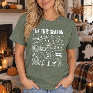 A woman wearing an olive green graphic t-shirt with white Halloween-themed illustrations and text reading 'TIS THIS SEASON'. The background shows a cozy, warmly lit living room with candles, pumpkins, and a fireplace.