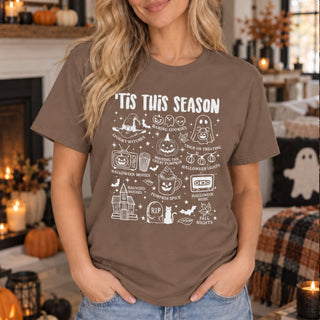 A woman wearing a brown graphic t-shirt with white Halloween-themed illustrations and text, standing in a cozy, decorated living room with candles and pumpkins.