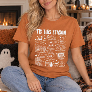 A woman wearing an orange crewneck t-shirt with a white graphic that reads 'TIS THIS SEASON' and features various Halloween-themed illustrations, sitting on a couch in a cozy living room setting.