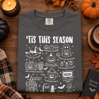 A dark gray t-shirt with a white 'Tis This Season' Halloween graphic print, laid flat on a rustic wooden table surrounded by seasonal props like a lit candle, a small pumpkin, and a plaid scarf.