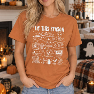 A woman wearing a burnt orange graphic t-shirt with white Halloween-themed illustrations and text reading 'TIS THIS SEASON'. The background shows a cozy, decorated fireplace with pumpkins, candles, and autumn decor.