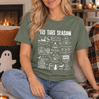 A woman sitting on a couch wearing an olive green graphic t-shirt with white Halloween-themed illustrations and text that reads 'IT'S THIS SEASON'.